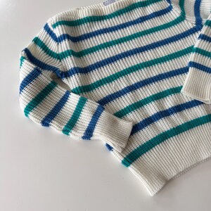 May include: A cream-colored sweater with horizontal stripes in teal and blue. The sweater has long sleeves and a ribbed hem and cuffs. The knitwear is folded on a white surface.