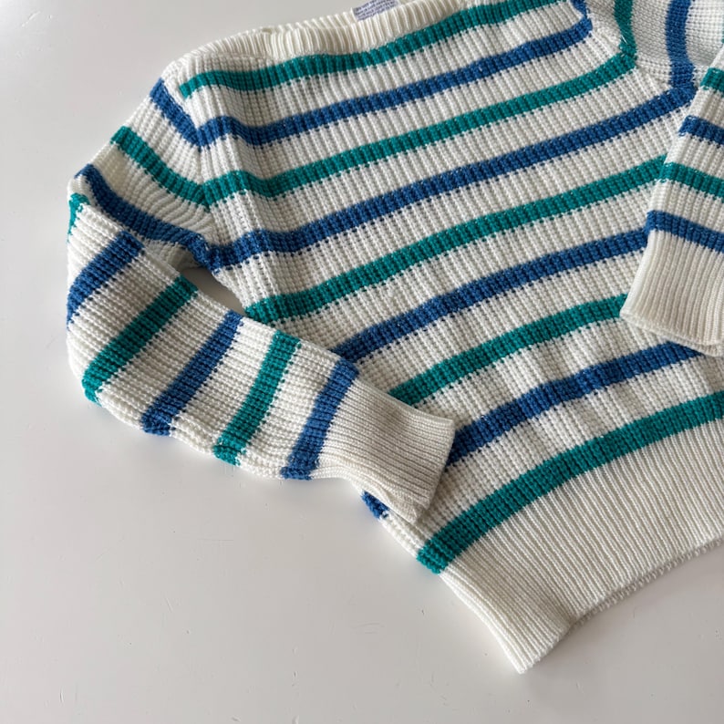 May include: A cream-colored sweater with horizontal stripes in teal and blue. The sweater has long sleeves and a ribbed hem and cuffs. The knitwear is folded on a white surface.