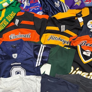 Peut inclure: Une collection de vêtements de sport assortis comprenant des maillots, des t-shirts et des shorts arborant divers logos et couleurs d'équipes tels que Minnesota, Notre Dame, Cincinnati, Pittsburgh, Dallas Cowboys et Winnipeg Jets, disposés en couches.