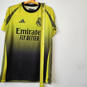 May include: A lime green and black gradient soccer jersey with a black crew neck, featuring the text Emirates FLY BETTER, a black logo, and a team crest. A yellow measuring tape is placed vertically along the front, showing a length of 22 inches.