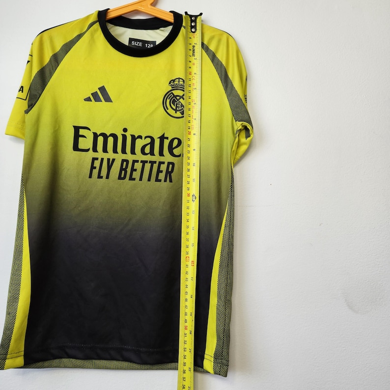 May include: A lime green and black gradient soccer jersey with a black crew neck, featuring the text Emirates FLY BETTER, a black logo, and a team crest. A yellow measuring tape is placed vertically along the front, showing a length of 22 inches.