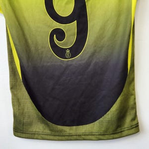 May include: A sports jersey featuring a lime green to black gradient design. The back displays a large black number nine with a small circular emblem featuring a crown and the letters RMCF positioned directly below the number. The sides have a textured pattern.