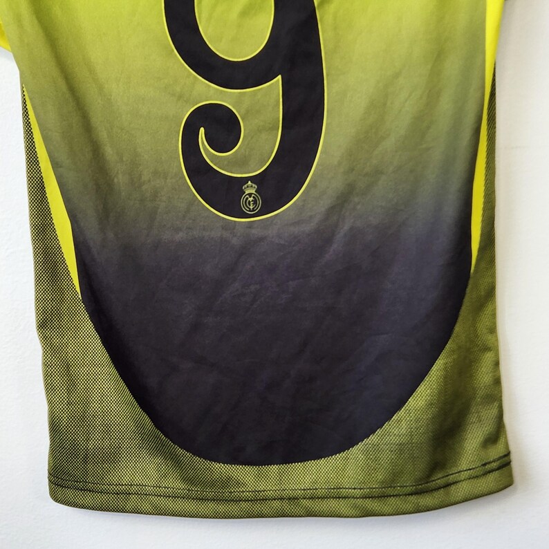 May include: A sports jersey featuring a lime green to black gradient design. The back displays a large black number nine with a small circular emblem featuring a crown and the letters RMCF positioned directly below the number. The sides have a textured pattern.