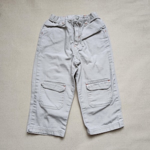 May include: Pale grey children's cargo trousers with two front pockets and orange stitching. The trousers feature a button closure and are made of a durable fabric. A casual style for everyday wear.