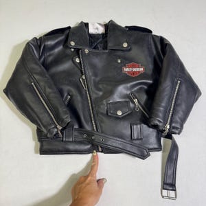 Könnte beinhalten: Eine schwarze Motorradjacke mit asymmetrischem silberfarbenem Reißverschluss, einem Taillengürtel mit Metallschnalle und Reißverschlussbündchen. Auf der Brust befindet sich ein rot-weiß-schwarzes Harley-Davidson-Logo. Sie hat eine kleine Druckknopftasche.