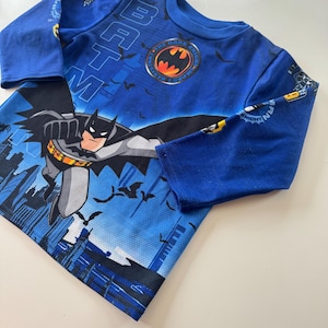 May include: Blue long-sleeved Batman shirt featuring a graphic of Batman flying over a cityscape. The shirt has the Batman logo on the chest and the word "Batman" printed on the sleeves.