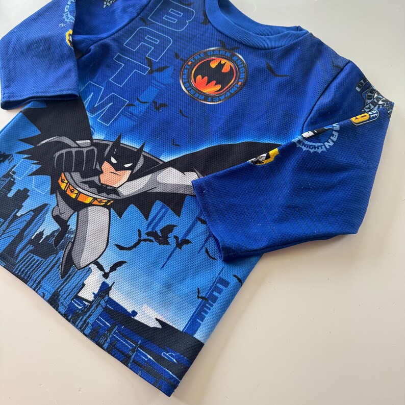 May include: Blue long-sleeved Batman shirt featuring a graphic of Batman flying over a cityscape. The shirt has the Batman logo on the chest and the word "Batman" printed on the sleeves.