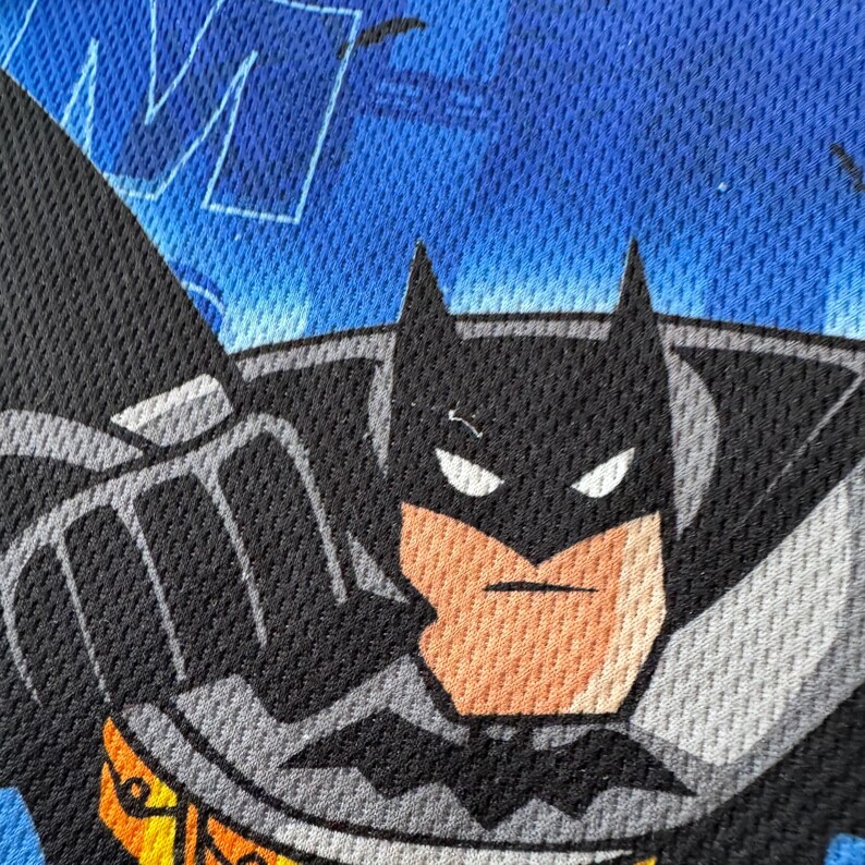 May include: Close-up of a Batman graphic on a blue and black mesh fabric. The Batman image features a black cowl, gray cape, and a yellow and black emblem. The background has a blue, textured pattern with white lettering.