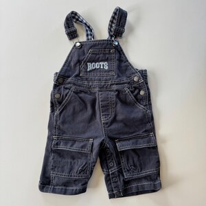 May include: Dark blue denim baby overalls with the word "ROOTS" embroidered on the chest pocket. The overalls have two front pockets, adjustable straps with a plaid pattern, and snap closures on the sides.