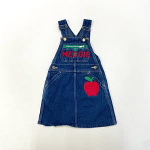 Peut inclure: Salopette en jean bleu avec une pomme rouge et le nom "MEGGIE" brodé sur le devant.