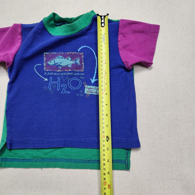 May include: A colorful short-sleeved t-shirt with a blue body, green hem, and purple sleeves. The shirt features a fish graphic and the text "H2O". A measuring tape shows the shirt is approximately 16 inches long.