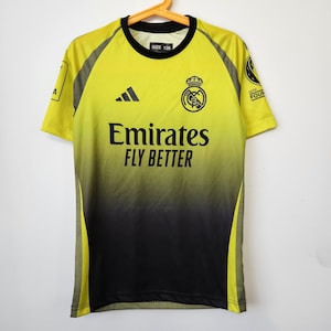 May include: A short-sleeve soccer jersey featuring a yellow-to-black gradient design. The shirt displays the Adidas logo, the Real Madrid crest, and the text Emirates Fly Better. A size 128 label is visible at the neck, with UEFA Foundation patches.