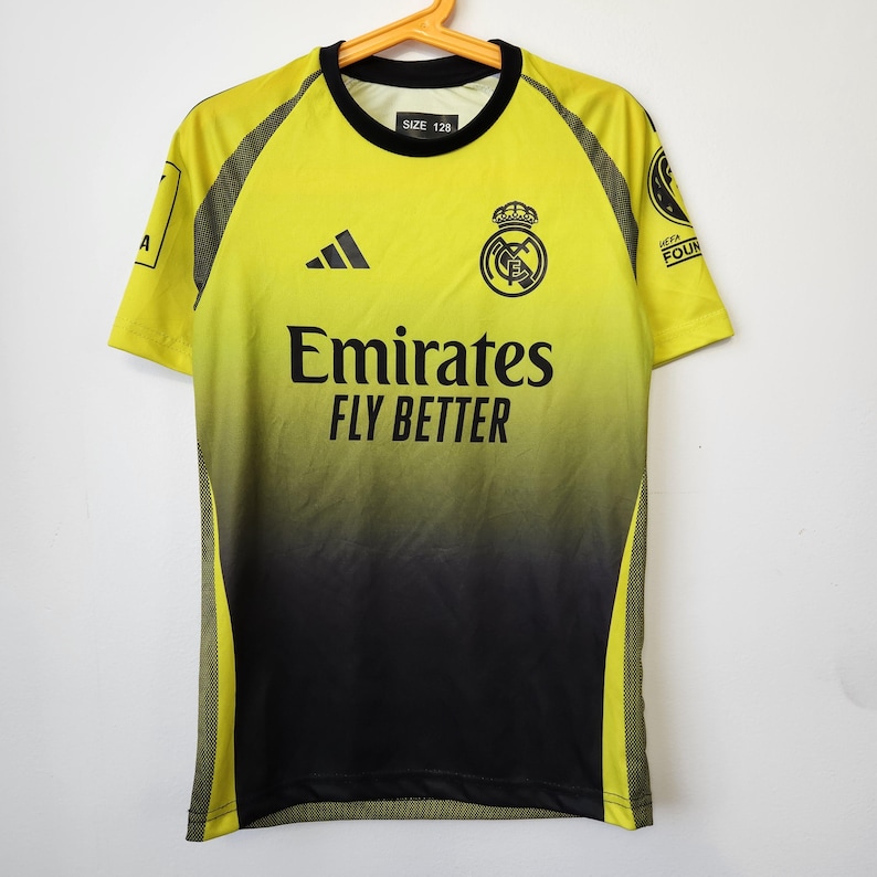 May include: A short-sleeve soccer jersey featuring a yellow-to-black gradient design. The shirt displays the Adidas logo, the Real Madrid crest, and the text Emirates Fly Better. A size 128 label is visible at the neck, with UEFA Foundation patches.