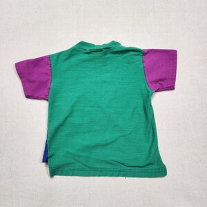 May include: A green and purple short-sleeved shirt. The shirt has a green body with horizontal ribbing and purple sleeves. A small section of blue fabric is visible on the side of the shirt.