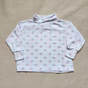 Könnte beinhalten: Weißes Rollkragenshirt mit einem Blumenmuster in Rosa und Blau. Das langärmlige Shirt ist aus weichem Material und für Kinder geeignet.