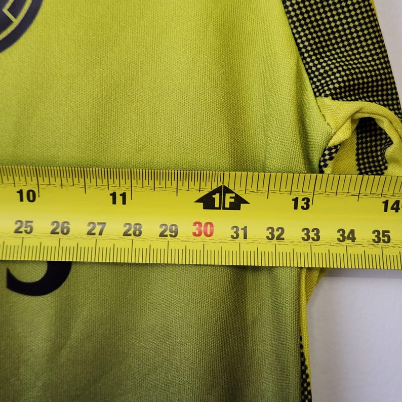 May include: A lime green shirt with black accents on the sleeve and a partial black graphic. A yellow measuring tape is placed across the chest, showing a width measurement of approximately 4.5 inches between the 10 and 14.5 inch marks.