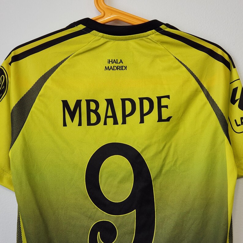 May include: A yellow and black soccer jersey featuring the name MBAPPE and the number 9 printed in black on the back. The text &iexcl;HALA MADRID! is printed in black near the neckline, with black mesh side panels and black shoulder stripes.