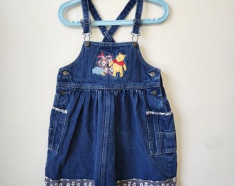 May include: A dark blue denim overall dress featuring adjustable shoulder straps with metal buckles. The chest displays an embroidered Winnie the Pooh character patch. The dress includes two side pockets and a hemline with a white floral pattern.