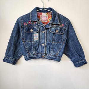 Peut inclure: Une veste en jean bleu délavé à l'acide avec un col, des manches longues et des boutons en métal. L'avant présente des broderies florales colorées, deux poches poitrine et un écusson rectangulaire. La doublure intérieure a un motif floral.