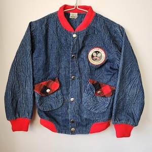 May include: A blue denim jacket with a crinkled texture, featuring a red ribbed collar, cuffs, and hem. It has a front button closure, two pockets with red and black plaid flaps, and a circular patch on the chest with the text SPORTS and tennis rackets.
