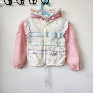May include: A white hooded jacket with long pink sleeves, featuring horizontal polka dot patterned stripes, small colorful embroidered dots, and three decorative bows on the right side. The jacket has a front zipper and a drawstring hem.