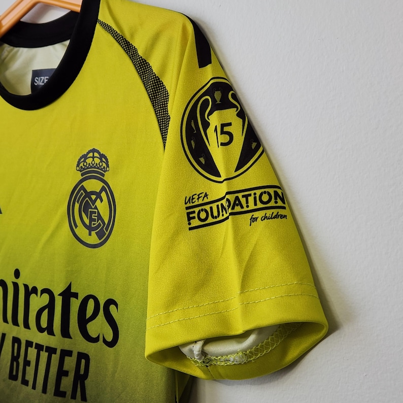 May include: A bright yellow short-sleeved jersey with a black crew neck collar. The front features the Real Madrid crest and the text Emirates Fly Better. The sleeve displays a black UEFA Foundation for children logo with the number 15 inside.