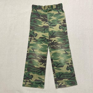 May include: Camouflage pants with a green, brown, and black pattern. The pants have a button and zipper closure and belt loops.