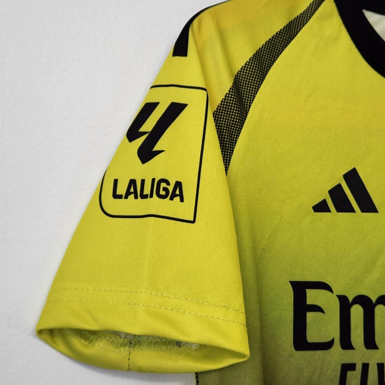 May include: A bright yellow short-sleeved sports jersey featuring a black Adidas logo and a black LaLiga patch on the sleeve. The fabric displays a gradient effect with black accents along the shoulder and collar area, including partial text.