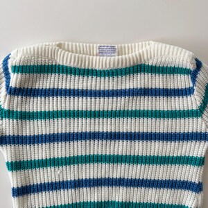 May include: A cream-colored sweater with horizontal stripes in teal and blue. The sweater has a ribbed crew neck and a tag at the back of the neck. The stripes are evenly spaced across the body of the sweater.
