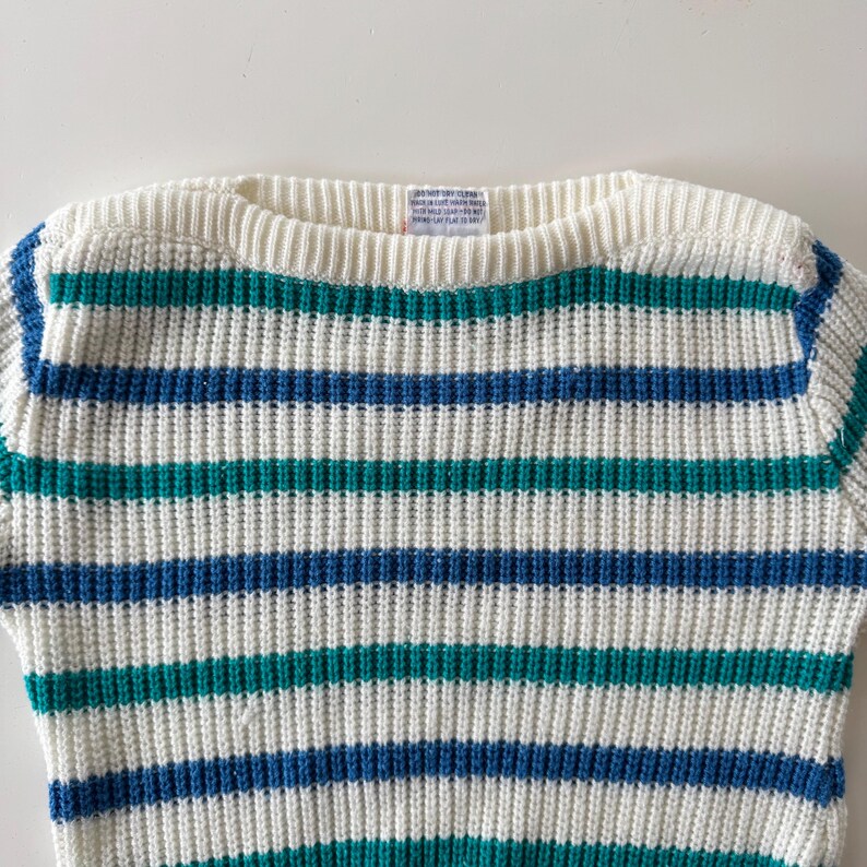 May include: A cream-colored sweater with horizontal stripes in teal and blue. The sweater has a ribbed crew neck and a tag at the back of the neck. The stripes are evenly spaced across the body of the sweater.