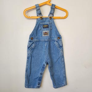 Può includere: Salopette in denim azzurro con spalline regolabili con fibbie in metallo, una tasca frontale sul petto con etichetta OshKosh B'gosh e una toppa rettangolare con la scritta New Directions sopra una grafica colorata. Le gambe hanno bottoni a pressione.