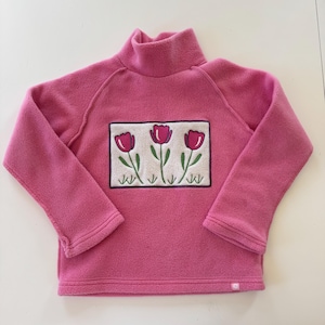 May include: A pink fleece turtleneck sweater with a floral design. The sweater features a rectangular patch with three red tulips with green stems and leaves. The sweater has long sleeves and a small logo near the bottom hem.