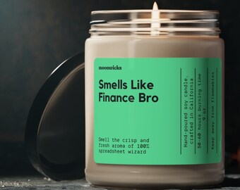 May include: A lit, clear glass jar candle with a green label that reads "Smells Like Finance Bro." The candle has a white wax and a black lid. The text on the label also says "Smell the crisp and fresh aroma of 100% spreadsheet wizard."