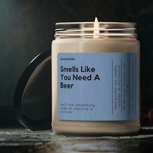 May include: A glass jar candle with a lit wick. The candle has a light blue label that reads "Smells Like You Need A Beer". The candle is hand-poured and crafted in California. The candle has a 60-hour burning time and weighs 9 oz.