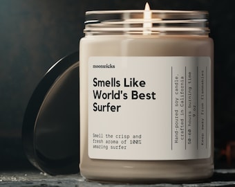 May include: A beige soy candle with a glass jar and a black lid. The candle is labeled "Smells Like World's Best Surfer" and has a description that says "Smell the crisp and fresh aroma of 100% amazing surfer". The candle is hand-poured and crafted in California. It has a 50-60 hour burn time and is 9 oz.