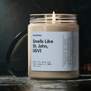 May include: A beige soy candle in a glass jar with a black lid. The candle is labeled "Smells Like St. John, USVI" and has a 50-60 hour burn time. The candle is hand-poured in California.