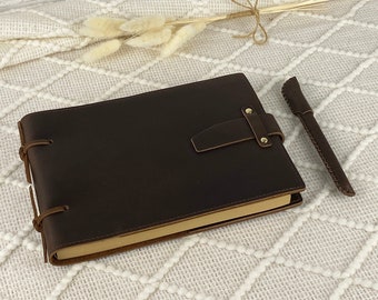 May include: A brown leather-bound journal with a strap closure and a matching brown leather pen. The journal is lying on a white textured surface.