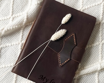 May include: A brown leather journal with the words "My love..." embossed on the cover. The journal is lying on a white knitted blanket with dried white flowers on top.