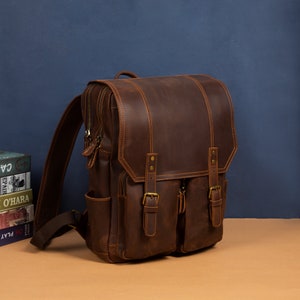 May include: A brown leather backpack with two front pockets and adjustable straps. The backpack has a distressed look and features brass buckles.