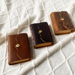 May include: Three small leather-bound journals with gold clasps. The journals are brown, black, and a light brown with an embossed design. The journals are on a white textured surface.
