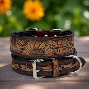 May include: Two stacked leather dog collars with sunflower and scrollwork designs. The collars are brown with black and burgundy accents, featuring silver-colored metal buckles and hardware. The collars are on a wooden surface with a blurred green background.