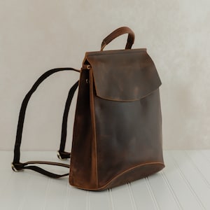 May include: A brown leather backpack with a flap closure and adjustable shoulder straps. The bag has a top handle and a minimalist design. The leather has a distressed finish, giving it a vintage look. The backpack is set against a neutral background.