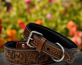 May include: Two brown leather dog collars with floral and geometric tooling. The collars have a black interior, silver-colored metal hardware, and white stitching. One collar is stacked on top of the other, with a blurred floral background.