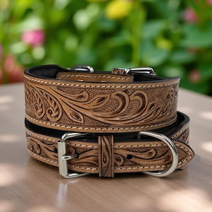 May include: Two stacked, light brown leather dog collars with intricate floral tooling. Each collar features a black interior, silver-toned metal hardware, and white stitching. The collars are designed for medium to large dogs.