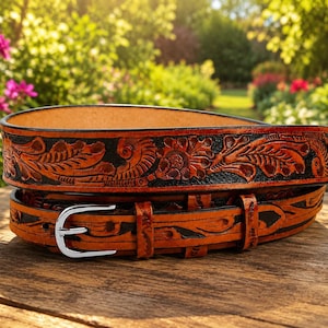 May include: A tan leather belt featuring a detailed floral and leaf pattern embossed into the surface with black dyed accents. The belt includes a polished silver-toned metal buckle and a matching leather keeper loop, coiled on a wooden surface.