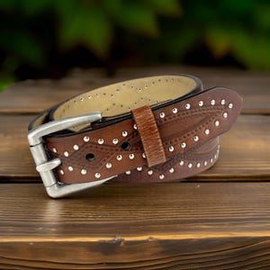 May include: A brown leather belt with silver studs and a decorative pattern. The belt has a silver buckle.