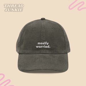 Op de afbeelding: Een donker olijfgroene corduroy baseballpet met de geborduurde witte tekst "mostly worried." gecentreerd op het voorpaneel. De pet heeft een gestructureerde kroon, een gebogen klep en bijpassende groene oogjes op de bovenste panelen.