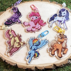 Puede incluir: Una colección de llaveros de dibujos animados de ponis coloridos. Cada llavero presenta un personaje de poni diferente con diseños y colores únicos, sujeto a un cierre de metal. Los llaveros se muestran sobre una rodaja de madera.