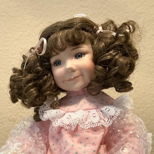 May include: A porcelain doll with brown curly hair, blue eyes, and a slight smile. The doll wears a pink dress with a white lace collar and small floral pattern. White ribbons adorn the doll's hair.