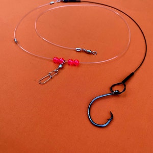 Puede incluir: Un aparejo de pesca con una línea transparente, cuentas rosadas y un anzuelo negro. El aparejo está unido a un giratorio.